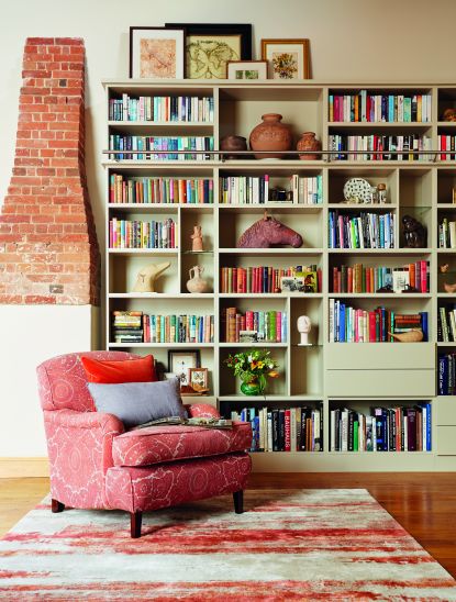 a luxurious spare room with a red armchair and rug, and a huge sage green built in bookcase unit with lots of books in it, and a brick covered-up fireplace next to it